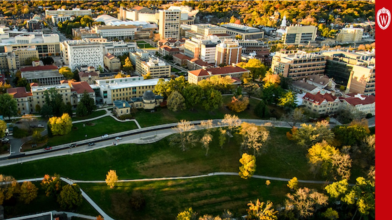 An aerial view of UW campus looks south from Observatory hill on a fall afternoon. Parkland in the foreground gives way to a cluster of academic buildings that span the width of the photo before transitioning to tree cover in the background.