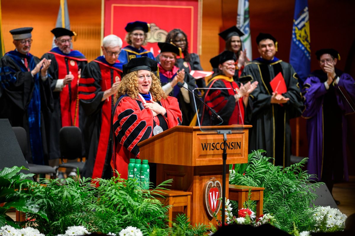 Jennifer Mnookin stands on stage with a crowd of dignataries, all wearing academic regalia. Mnookin folds her hands on her heart in a gesture of gratitude as she makes an address from a wooden podium with the word "Wisconsin" and the UW-Madison crest on the front.