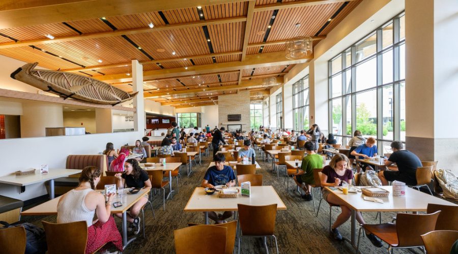 Students sit at tables in a cafeteria eating, with large windows facing them.
