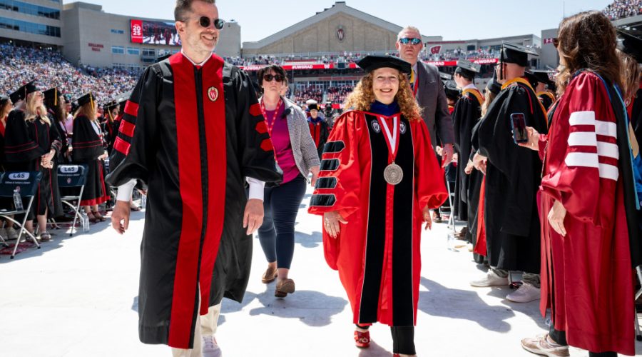 A number of people in academic robes walk down the aisle of a stadium.