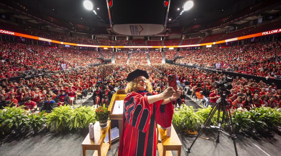 Chancellor Mnookin stands on stage, holding a phone as she takes a selfie of herself and the freshman class.