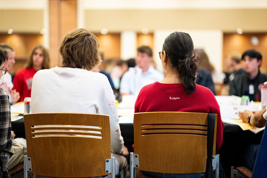 Two people with backs facing the camera sit at large table in conversation with other people