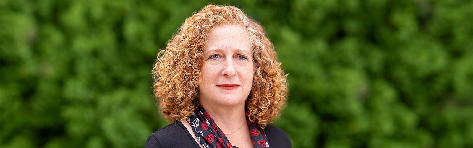 Chancellor Mnookin poses outdoors in front of a lush green background, wearing a dark top and a patterned scarf.