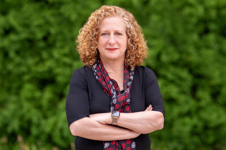 Chancellor Mnookin poses outdoors in front of a lush green background, wearing a dark top and a patterned scarf.