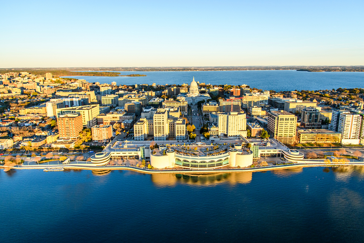 Aerial view of Madison, Wisconsin, showing the State Capitol centered on the isthmus between two lakes, with surrounding buildings in sunlight.