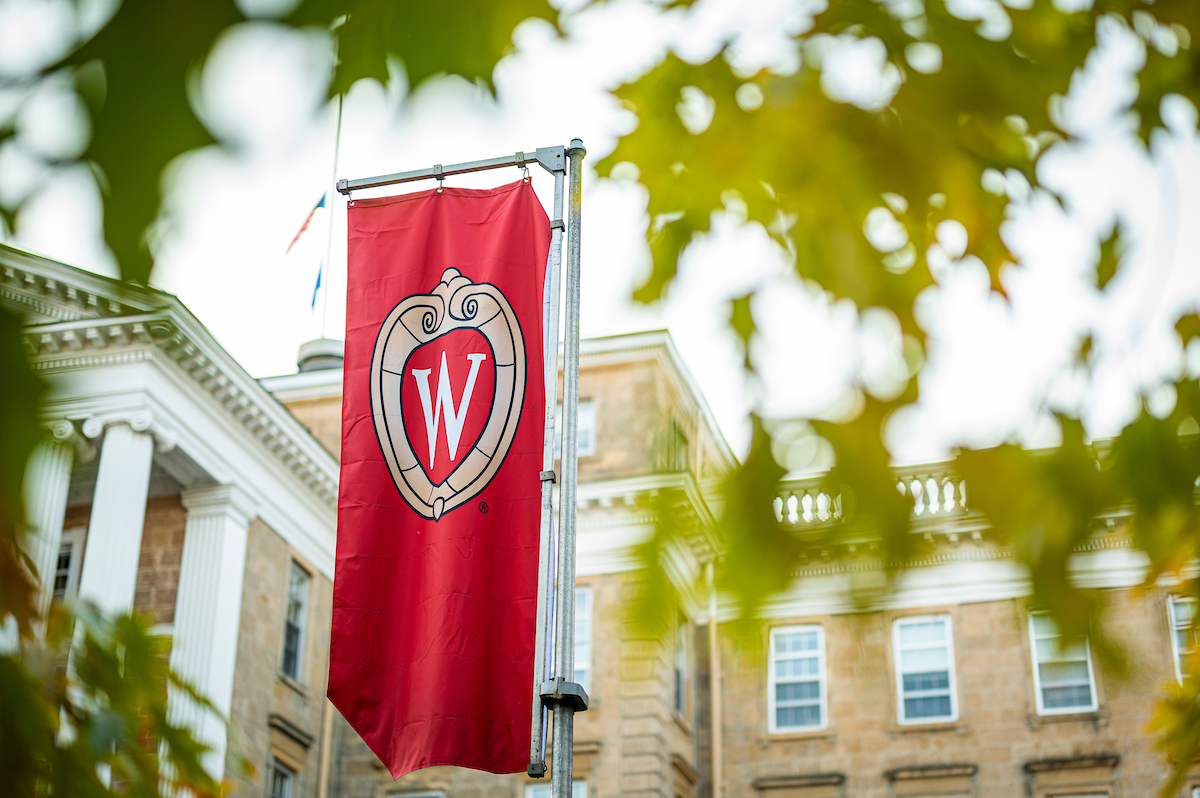 A red W crest banner positioned in front of Bascom Hall set among green leaves.