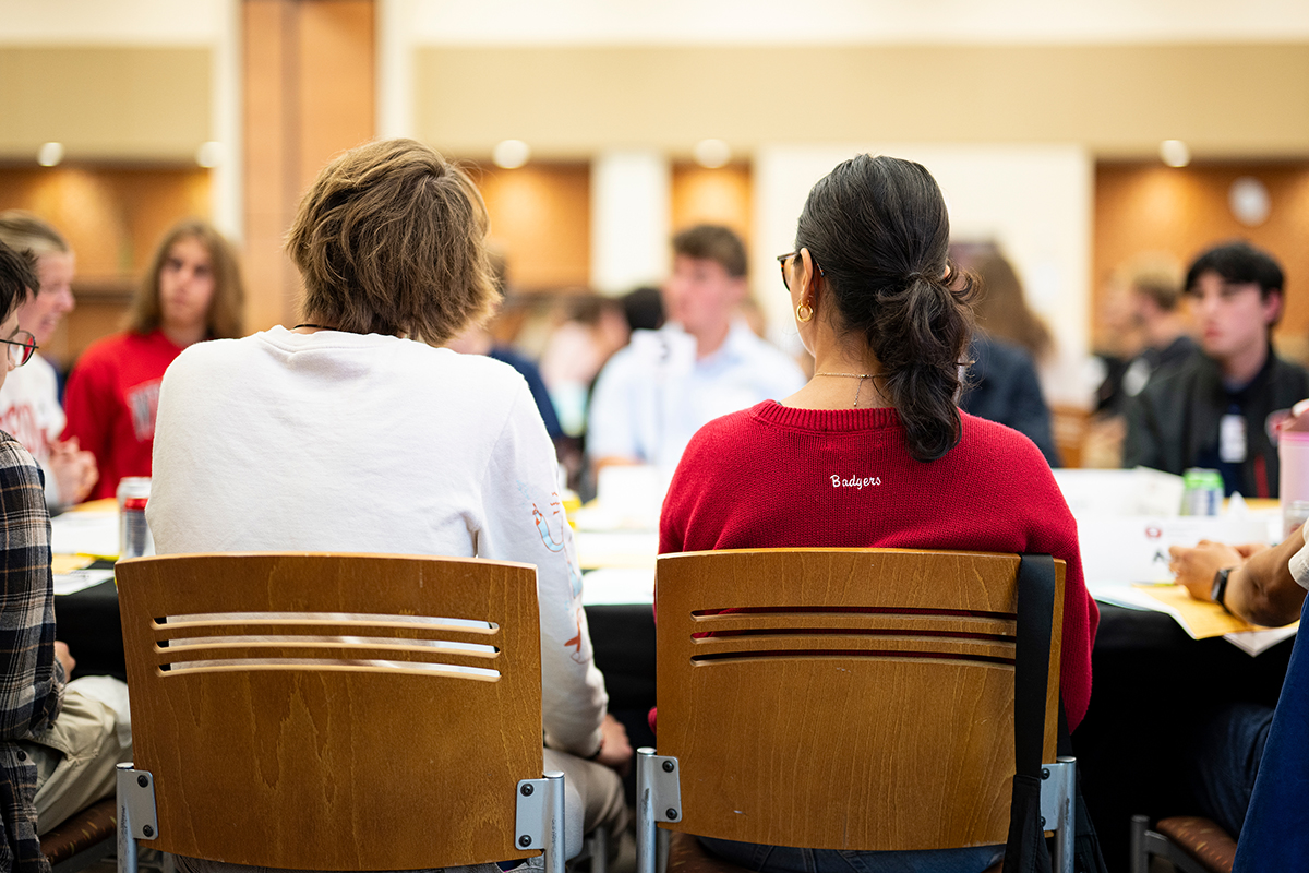People sit around a table in a workshop setting, with two individuals in the foreground viewed from behind, including one wearing a red “Badgers” sweater.