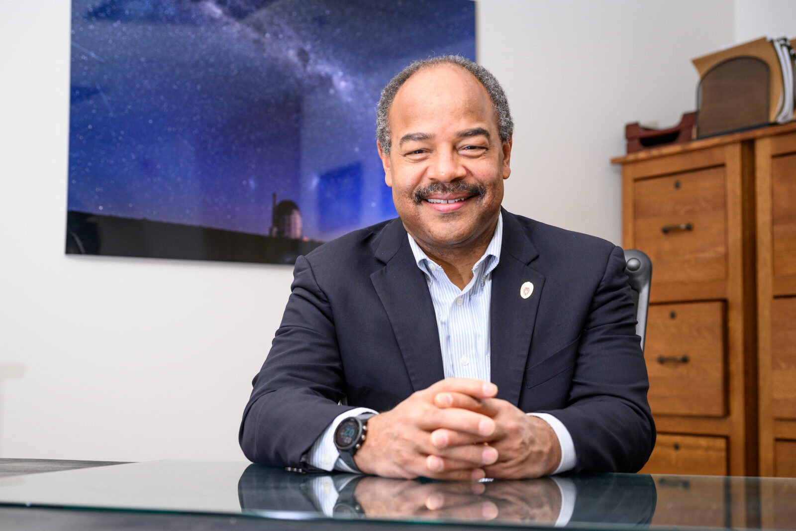 Eric Wilcots seated at a glass desk, hands clasped, with a starry‑sky photo behind him.