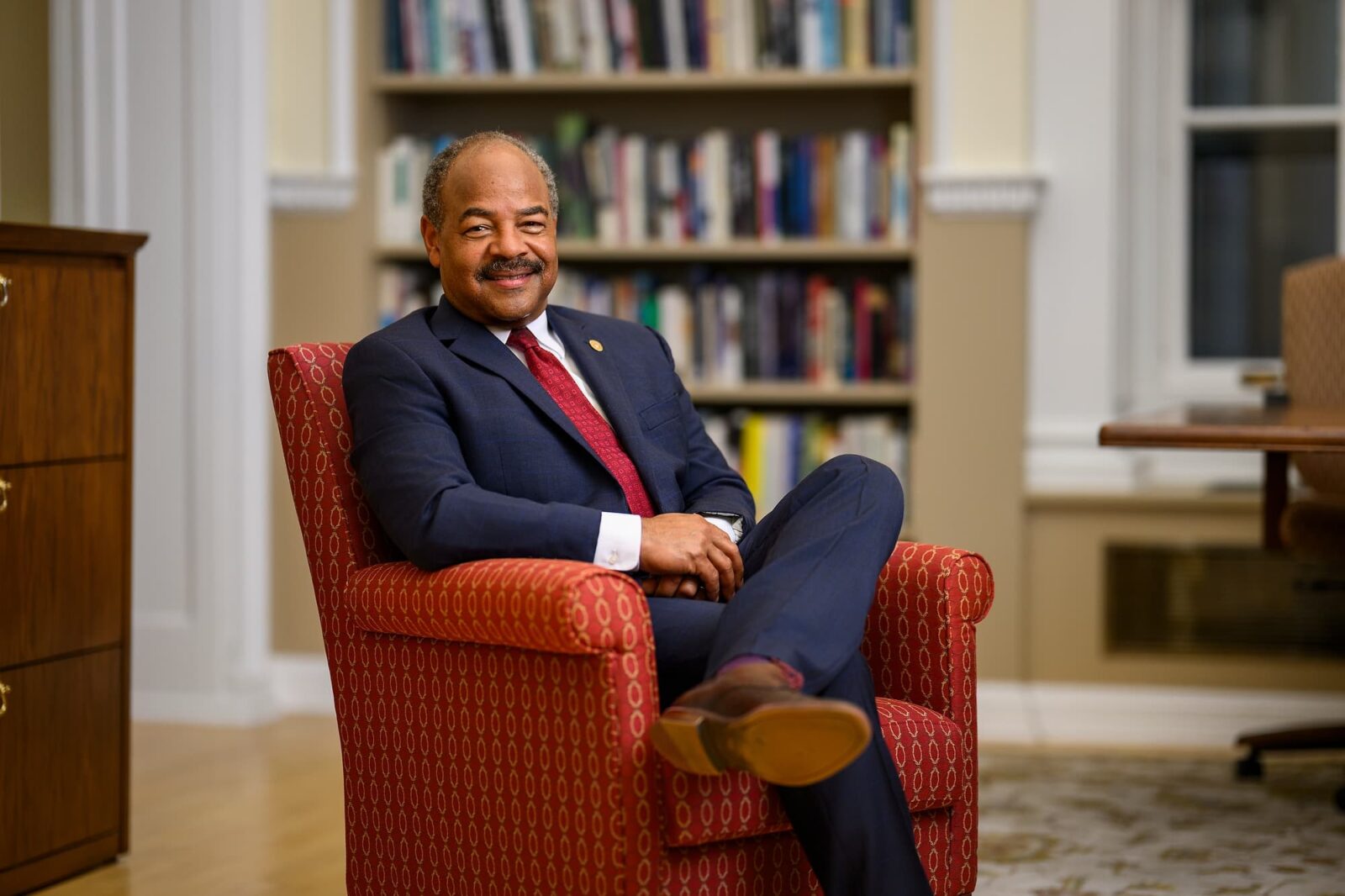 Eric Wilcots seated in a red armchair, wearing a navy suit and red tie, smiling in an office.