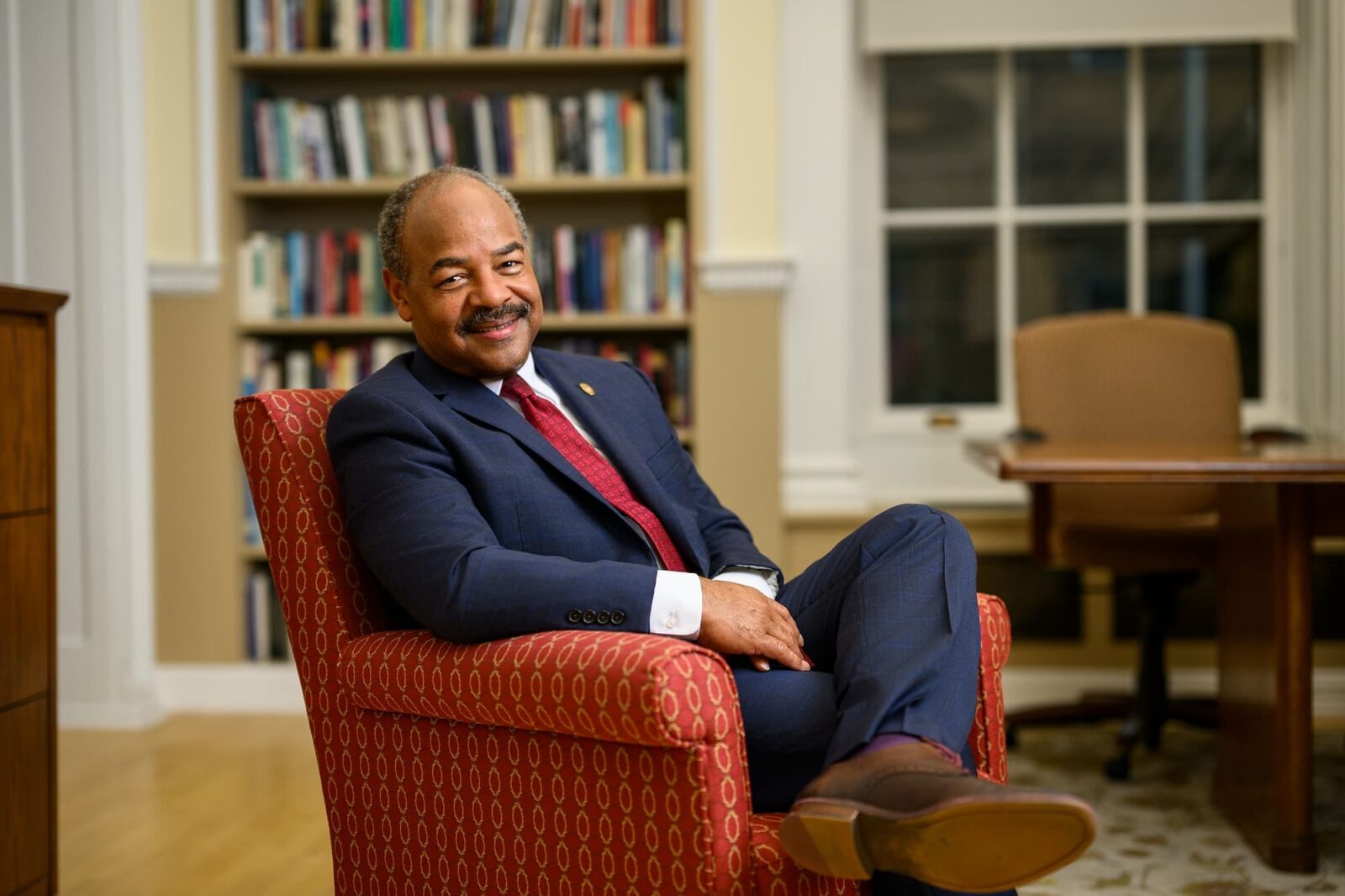 Eric Wilcots seated in a red armchair, wearing a navy suit and red tie, smiling in an office.