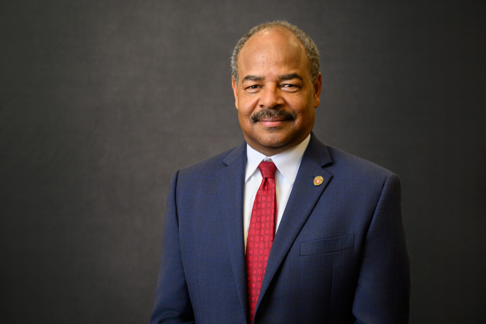 Portrait of Eric Wilcots wearing a navy suit and red tie against a gray backdrop. 
