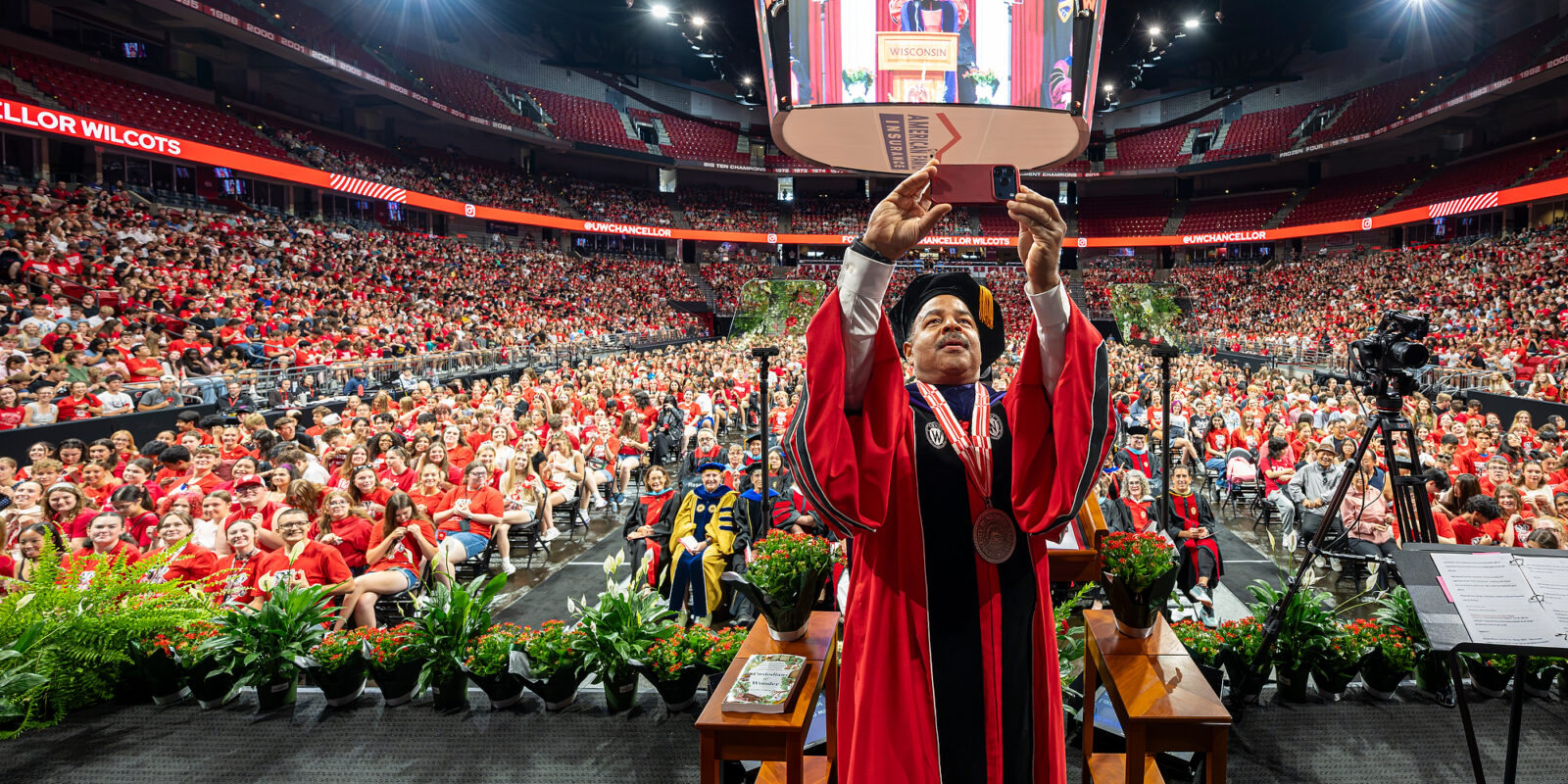 Interim Chancellor Wilcots holds up his phone and takes a selfie of himself and the thousands of students standing in the Kohl Center behind him.