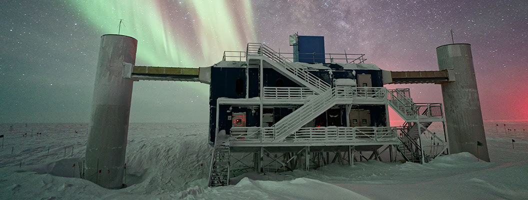 A view of the IceCube Lab with a starry night sky showing the Milky Way and light green and pink auroras.